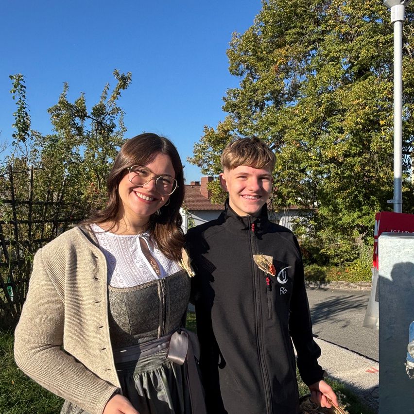 Eine Frau in einem traditionellen Kleid und ein Mann in einem schwarzen Jacke stehen in einem Park, lächeln und halten Körbe voller Pilze.