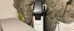 Ein medizinisches Gerät, mit der Marke 'esthetec', steht in einem Raum gegen eine Steinwand. Es verfügt über einen Monitor und ist an eine Steckdose angeschlossen. Ein Bett befindet sich in der Nähe.