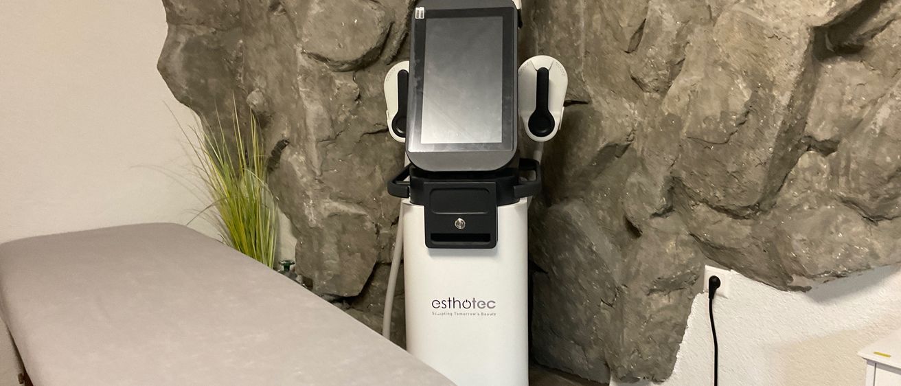 Ein medizinisches Gerät, mit der Marke 'esthetec', steht in einem Raum gegen eine Steinwand. Es verfügt über einen Monitor und ist an eine Steckdose angeschlossen. Ein Bett befindet sich in der Nähe.