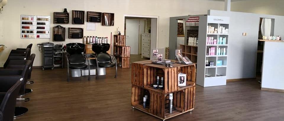 Ein leerer Salon mit Holzböden, weißen Wänden und verschiedenen Regalen und Schränken. Es gibt zwei schwarze Stühle und eine Waschstation mit zwei Waschbecken.