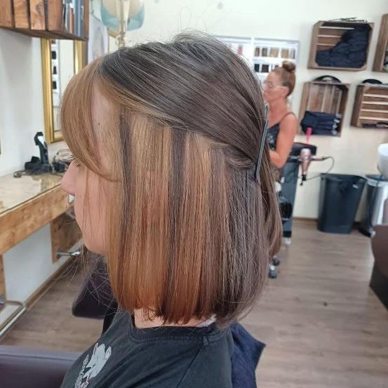 Eine Frau mit kurzen Haaren sitzt in einem Salon, ihr Haar mit Highlights und einem Zopf gestylt. Im Hintergrund arbeitet eine Frau mit einem Föhn.