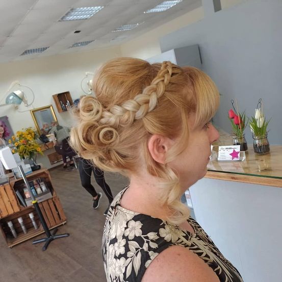 Eine Frau mit blondem Haar in einer Zopf- und Lockenfrisur steht in einem Salon und schaut auf eine Theke mit Blumen und einem Schild.