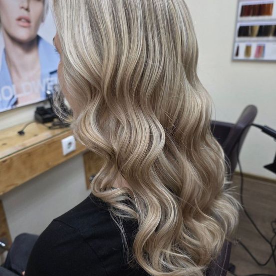 Eine Frau mit langen, welligen blonden Haaren sitzt in einem Salon und schaut in einen Spiegel, hinter dem ein Poster hängt.