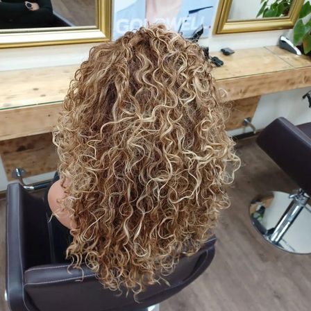 Eine Frau mit lockigem blondem Haar sitzt in einem Salonstuhl, dem Kameramann den Rücken zugewandt. Hinter ihr ist ein Holzschreibtisch und zwei goldgerahmte Spiegel zu sehen.