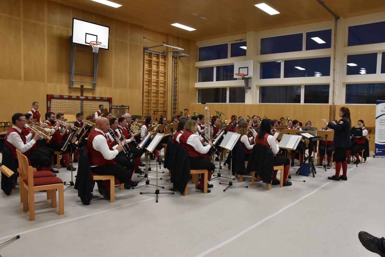Ein Blechbläserensemble spielt in einer Halle mit großen Fenstern, Basketballkörben und Holzwänden. Die Musiker sitzen und spielen verschiedene Instrumente.
