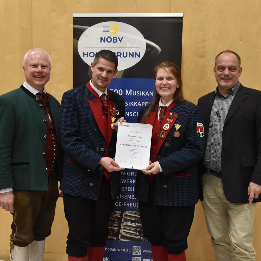 Vier Personen stehen nebeneinander. Eine Frau in der Mitte hält ein Zertifikat. Hinter ihnen ist ein Banner mit den Texten 'NOBV' und '100 Musiker'.