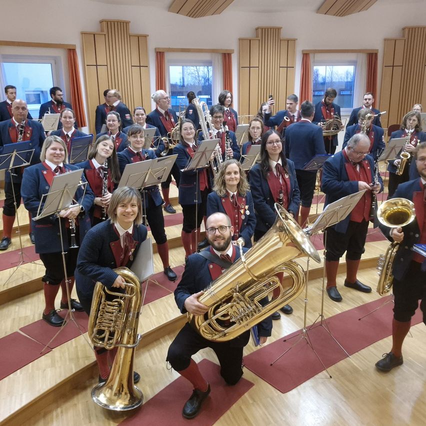 Ein Brass-Band steht auf der Bühne, wobei einige Mitglieder vorn Posaunen halten und andere dahinter verschiedene Instrumente. Die Umgebung ist drinnen mit Holzböden und einer Kulisse von Fenstern.