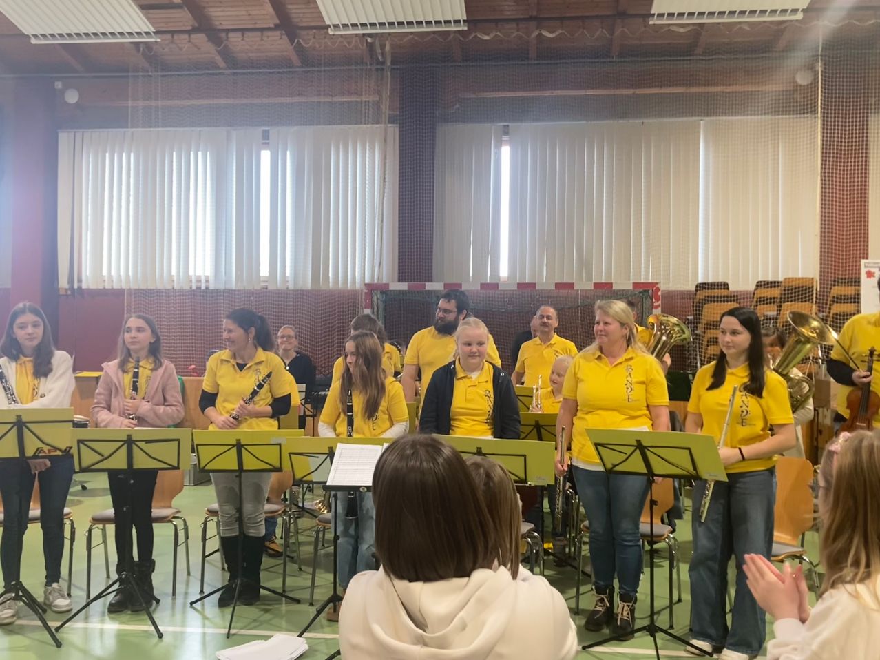 Eine Gruppe von Menschen in gelben Uniformen steht in einer Turnhalle, hält Musikinstrumente und übt. Eine Frau in einer weißen Kapuzenpulli beobachtet sie.
