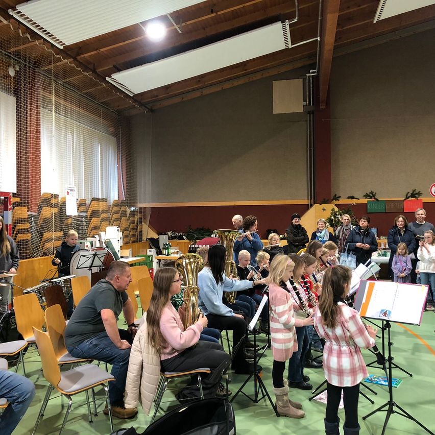 Ein Band-Probe in einem großen Innenraum. Mehrere Kinder spielen Instrumente, während andere zusehen, und Erwachsene sitzen auf Stühlen. Der Raum hat Holzbalken, weiße Decken und einen grünen Boden.
