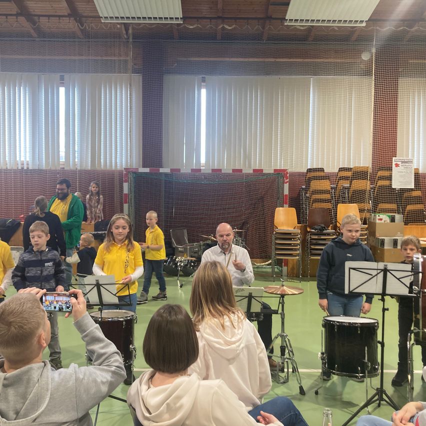 Mehrere Kinder in gelben Uniformen spielen in einem Raum Trommeln. Ein Mann sitzt auf einem Stuhl und spielt die Trommeln. Einige Leute sitzen auf Stühlen an der Seite. Dahinter sind gestapelte Stühle und ein Netz an der Wand. Es gibt ein Schild an der Wand.