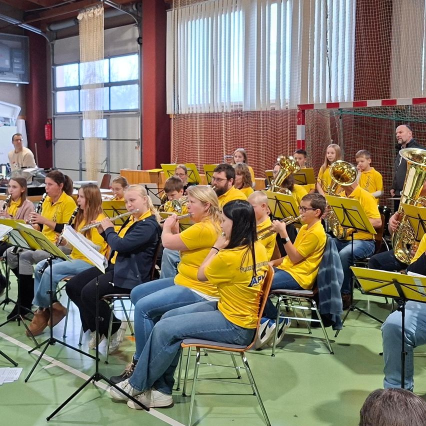 Eine Gruppe von Schülern in gelben Shirts übt mit ihren Instrumenten in einer Turnhalle. Sie sitzen auf Stühlen und spielen Trompeten, Posaunen und Flöten. Ein Mann steht in der Nähe eines Netzes im Hintergrund.
