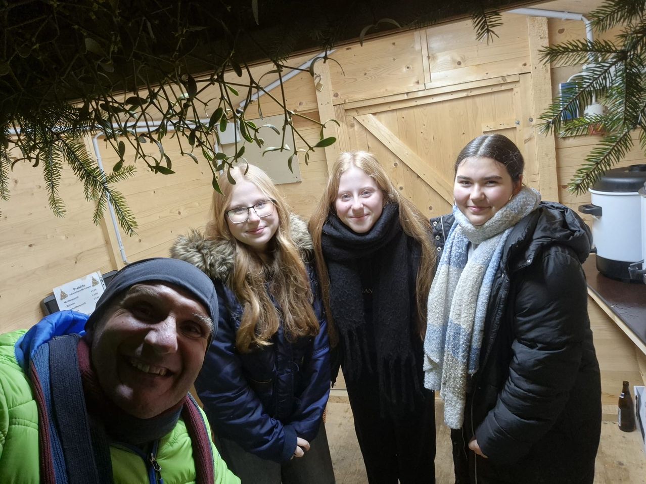 Eine Gruppe von vier Personen steht eng beieinander in einem hölzernen Raum und posiert lächelnd für ein Foto. Die beiden Frauen vorn tragen Brillen, Schals und Wintermäntel. Der Mann links trägt eine Mütze und eine Jacke.