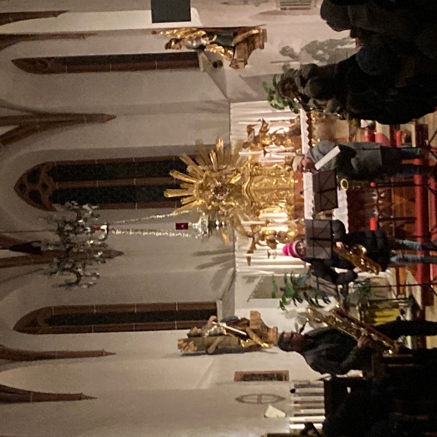 Eine Gruppe von Musikern tritt in einer Kirche auf. Goldene Engelsstatuen und eine Kristallkronleuchter hängen von der Decke. Das Publikum steht und schaut zu.
