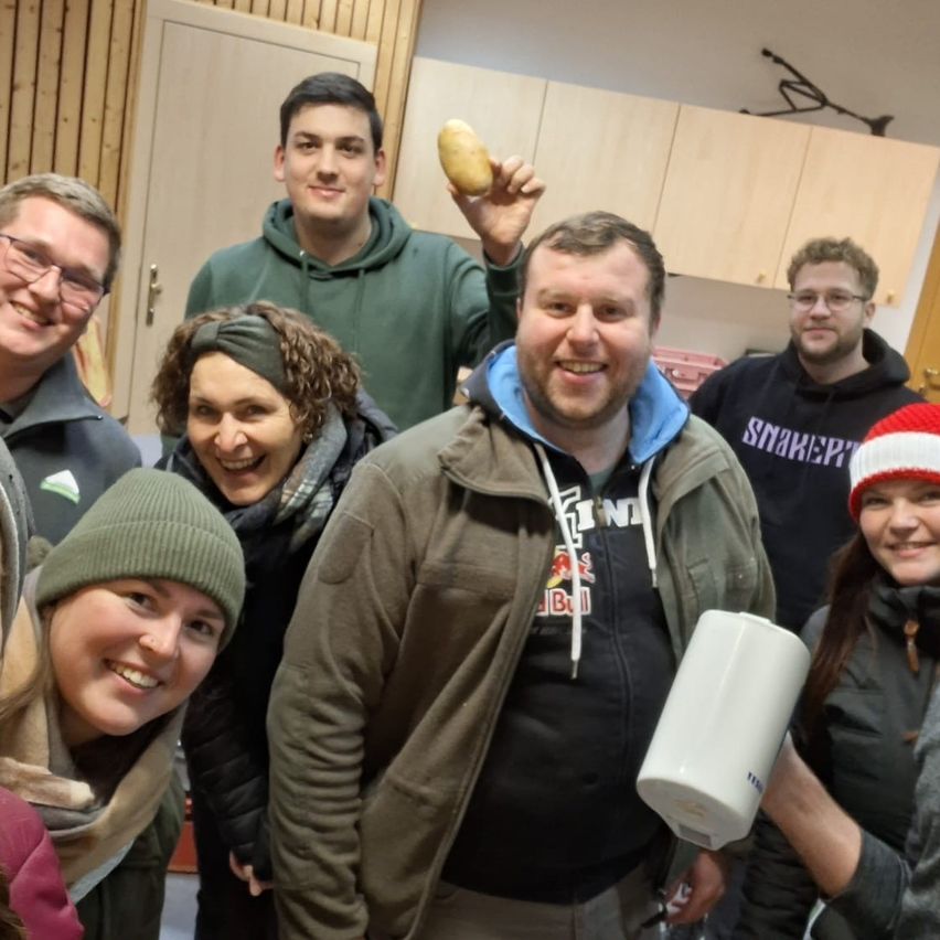 Eine Gruppe lächelnder Erwachsener in Winterkleidung posiert für ein Foto in einem Raum. Ein Mann hält eine Kartoffel und ein anderer eine weiße Tasse.