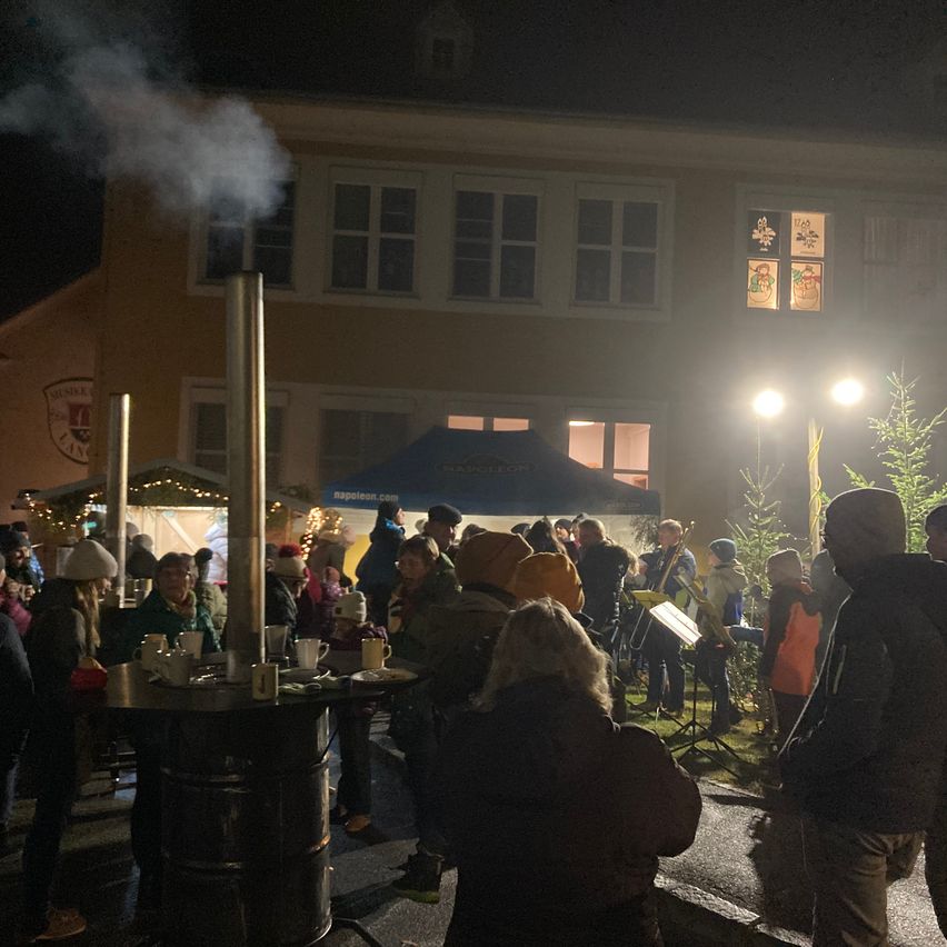 Eine Menschenmenge versammelt sich um einen Tisch mit Tassen und Tellern auf einem nächtlichen Markt unter einem blauen Zelt. Ein Schornstein lässt Rauch vor einem Gebäude mit beleuchteten Fenstern aufsteigen.