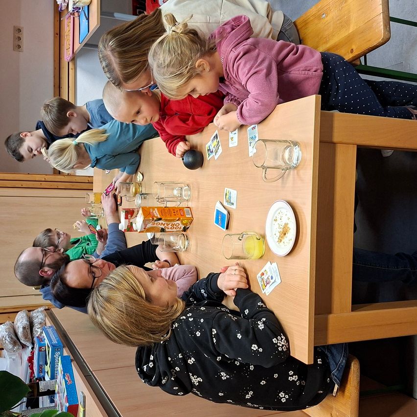 Eine Blick von oben auf Kinder, die um einen Tisch herum versammelt sind, einige lehnen sich vor, andere sitzen, und beschäftigen sich mit Aktivitäten. Gläser, Karten und Essen sind auf dem Tisch. Erwachsene sitzen an der Seite, und eine Pflanze ist im Vordergrund.