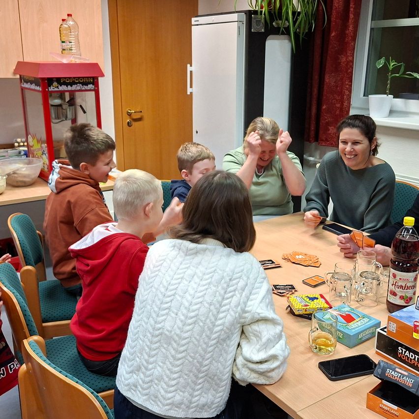 Eine Gruppe von Kindern und Erwachsenen sitzt um einen Tisch in einem Raum. Sie spielen ein Brettspiel und lachen.