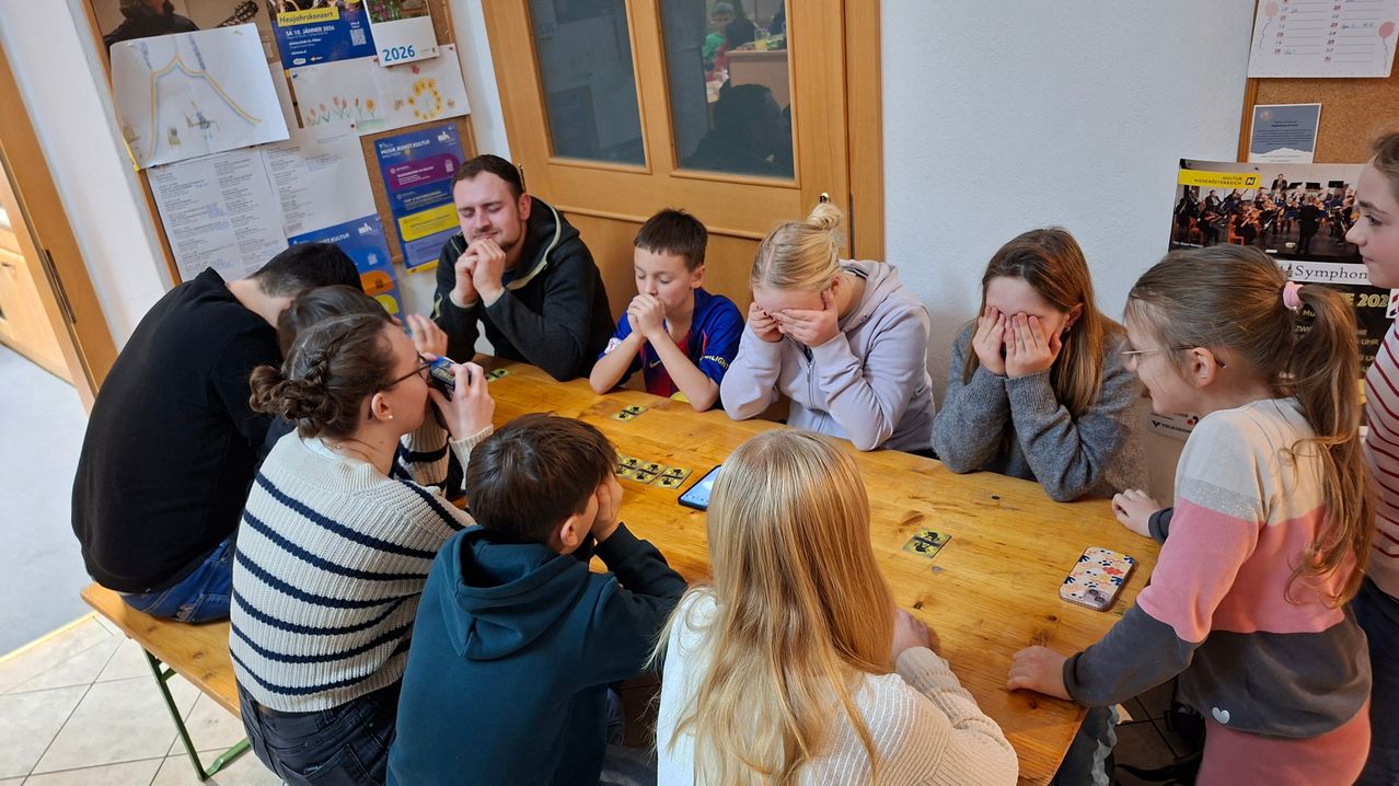 Eine Gruppe von Kindern und Erwachsenen sitzt um einen Holztisch herum, alle mit den Händen vor den Gesichtern. Ein Junge hält ein Handy. Der Tisch hat Karten darauf.