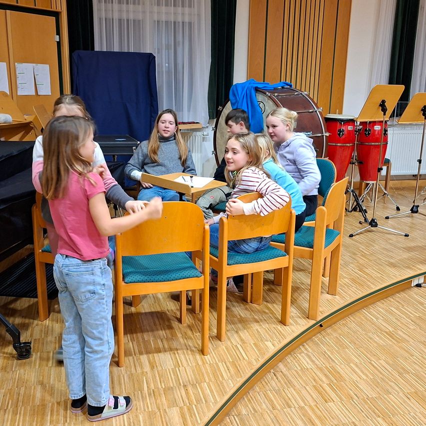 Eine Gruppe junger Mädchen versammelt sich in einem Raum, sitzt um einen Tisch herum, mit Musikinstrumenten im Hintergrund.