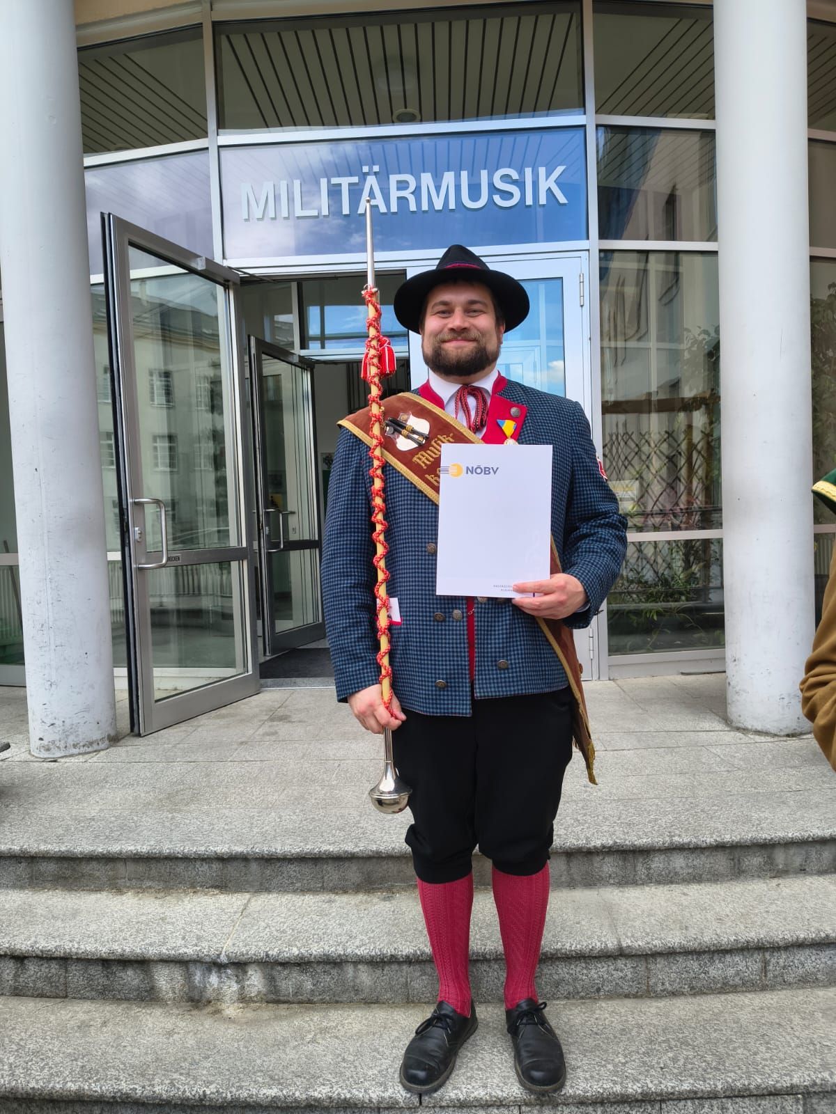 Ein Mann in traditioneller Kleidung steht vor einem Gebäude mit einer Trompete und einem Papier. Er lächelt und hält die Trompete in einer Hand. Das Gebäude hat eine Glastür mit 'MILITÄRMUSIK' darauf.