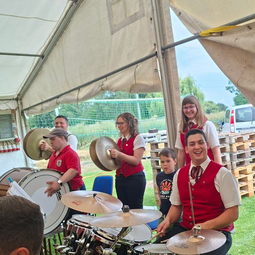 Eine Gruppe von Menschen in roten Westen und Hüten spielt Trommeln und Becken unter einem Zelt. Ein kleiner Junge steht in der Nähe, und eine Frau lächelt im Hintergrund.