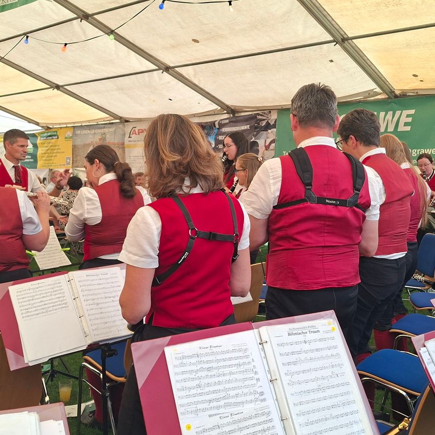 Eine Gruppe von Musikern in roten Westen und weißen Hemden steht in einem Zelt, möglicherweise in Vorbereitung auf eine Aufführung. Sie halten Notenblätter in den Händen und einige tragen Brillen.