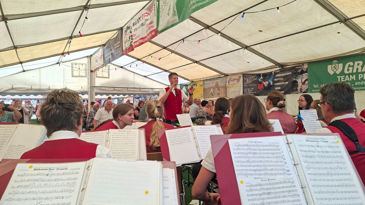 Ein Mann in einer roten Weste dirigiert eine Band unter einem Zelt, während mehrere Menschen sitzen und zuhören. Die Bandmitglieder halten Notenblätter.