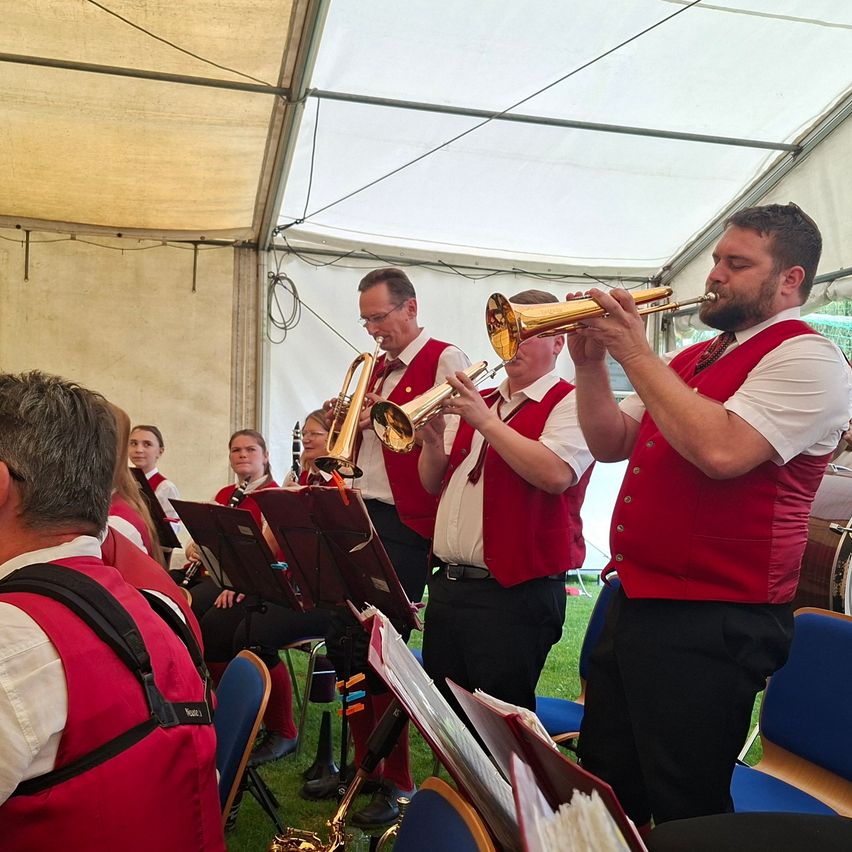 Eine Gruppe von Musikern in roten Westen und schwarzen Hosen spielt Trompeten und Posaunen unter einem Zelt. Einige sitzen, andere stehen.
