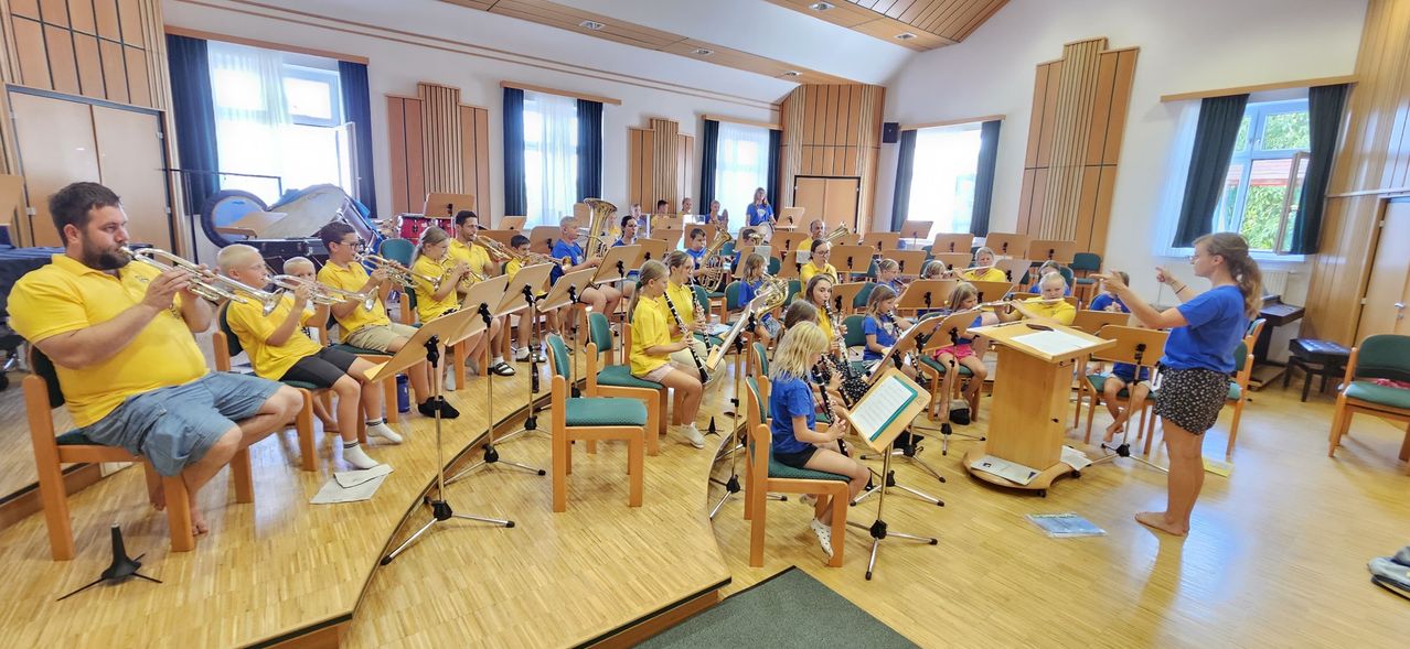 Eine Gruppe von Kindern in gelben und blauen Shirts sitzt auf Stühlen und spielt Musikinstrumente in einem Raum.