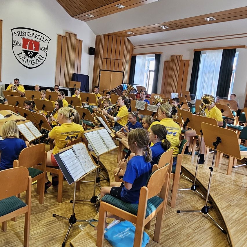 Ein Schulband-Probe in einem Raum mit Holzboden und einer Wand, die das Musikkapelle Langau-Logo zeigt. Schüler in gelben und blauen Shirts spielen verschiedene Instrumente, während sie auf Stühlen sitzen.