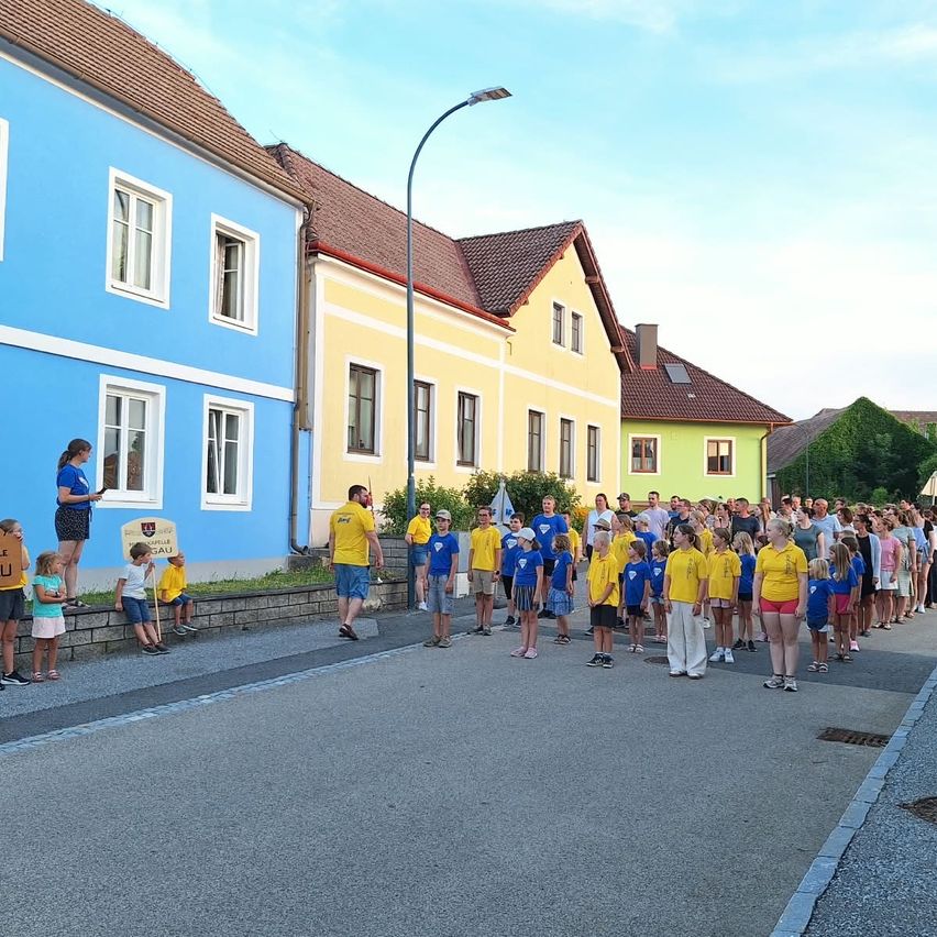Eine Gruppe von Kindern und Erwachsenen in gelben T-Shirts steht auf der Straße vor bunten Häusern. Eine Frau steht auf einer Mauer und hält ein Schild.