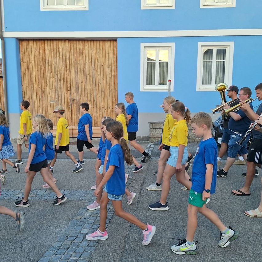 Eine Gruppe von Kindern in blauen Shirts und Shorts geht in einer Reihe vor einem blauen Gebäude mit einer Holztür und Fenstern. Erwachsene in blauen und gelben Shirts spielen Musikinstrumente.