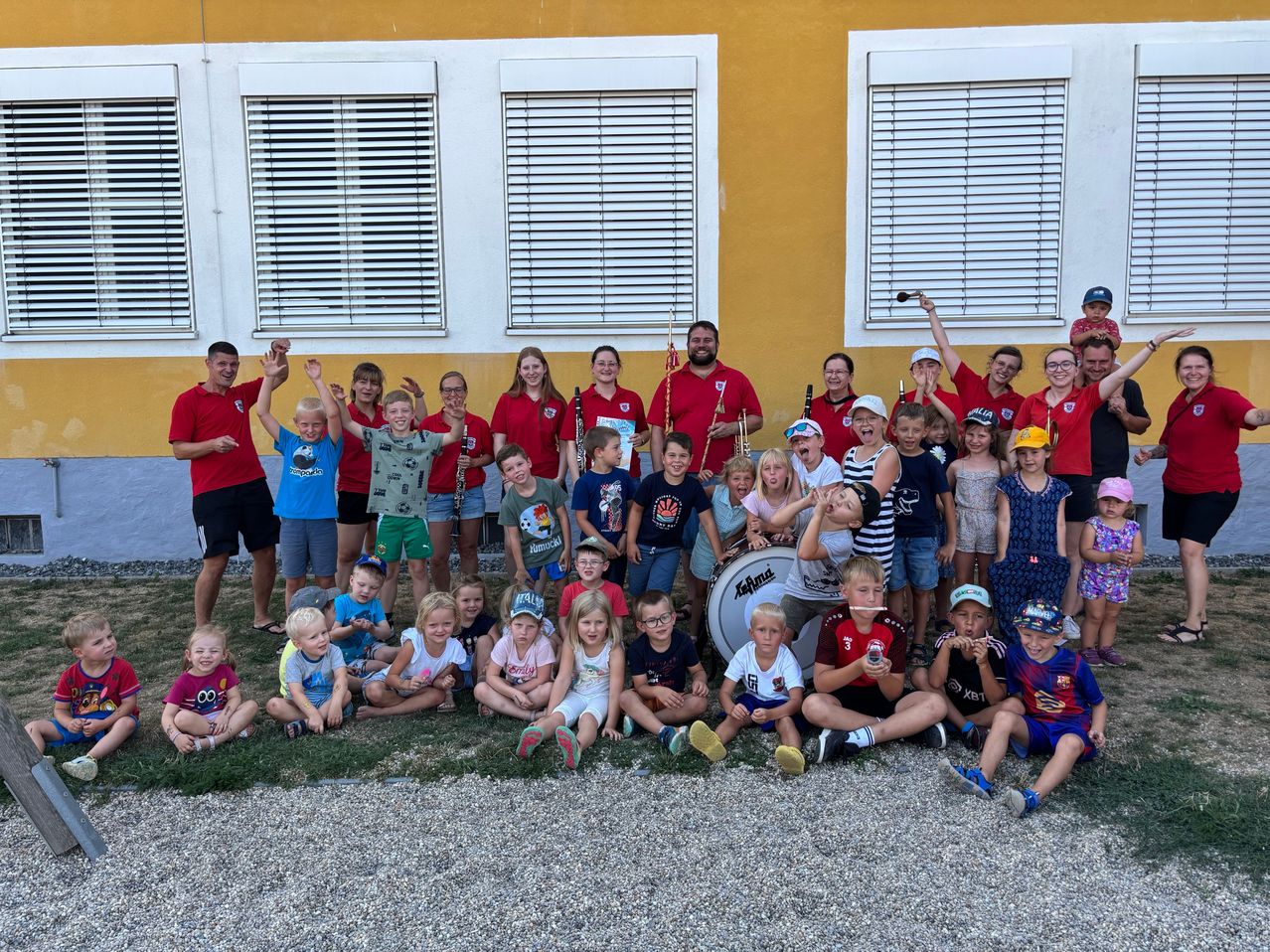 Eine Gruppe von Kindern und Erwachsenen in roten Shirts posiert für ein Foto vor einem gelben Gebäude mit weißen Fensterläden. Einige halten Musikinstrumente.