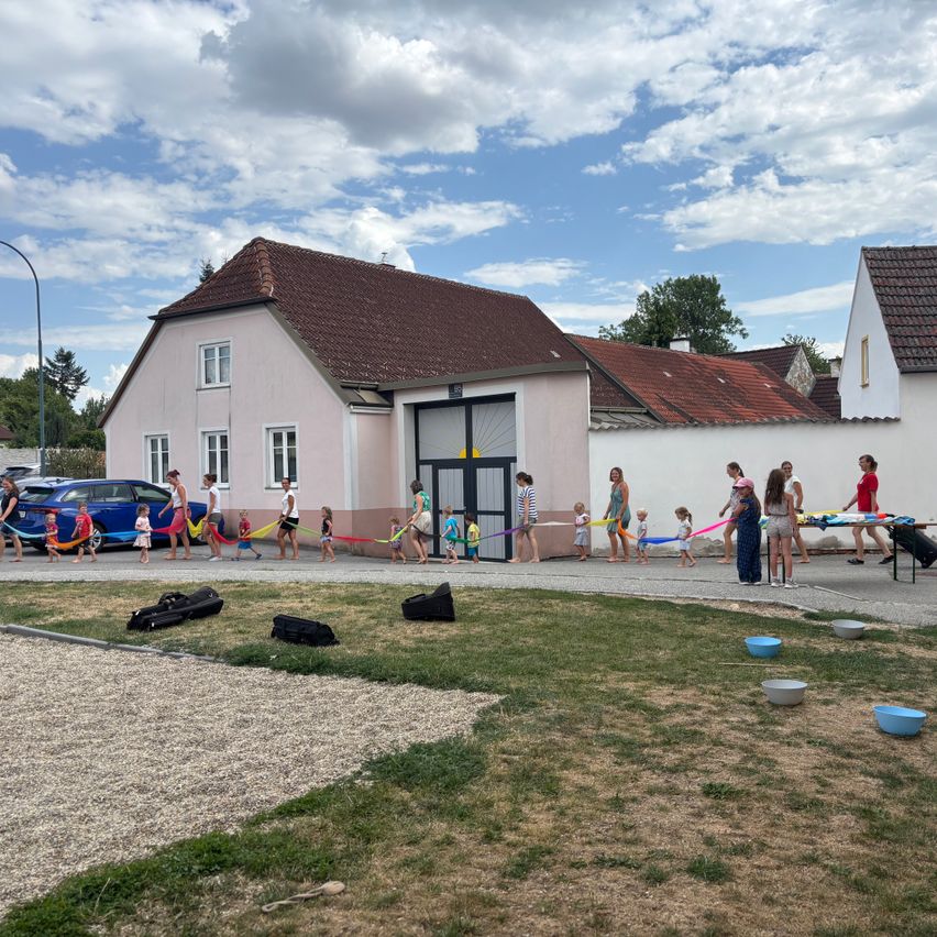 Eine Gruppe von Kindern und Erwachsenen hält bunte Bänder vor einem Haus mit braunem Dach an einem sonnigen Tag.