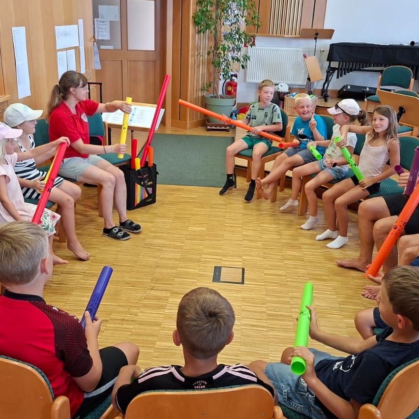 Eine Gruppe von Kindern und Erwachsenen sitzt im Kreis und hält bunte Musikinstrumente, möglicherweise Flöten, während eine Frau in der Mitte ein gelbes Instrument hält.