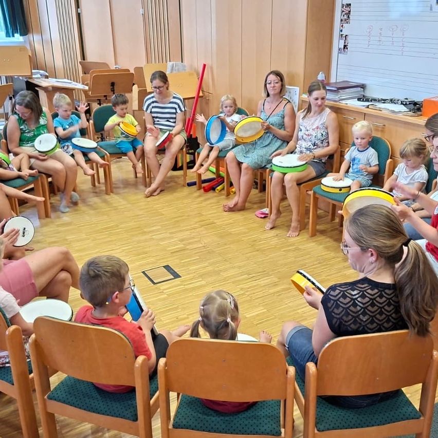 Eine Gruppe von Kindern und Erwachsenen sitzt im Kreis und spielt Trommeln und andere Percussionsinstrumente. Die Kinder halten Trommeln und einige spielen, während andere zuhören. Die Erwachsenen sitzen auf Stühlen und beobachten die Kinder. Der Raum hat Holzböden und Wände.