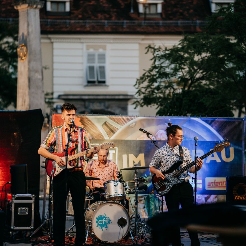 Eine Band spielt im Freien auf der Bühne mit einem großen Banner im Hintergrund, einer spielt Gitarre, einer spielt Schlagzeug und einer spielt Bassgitarre.
