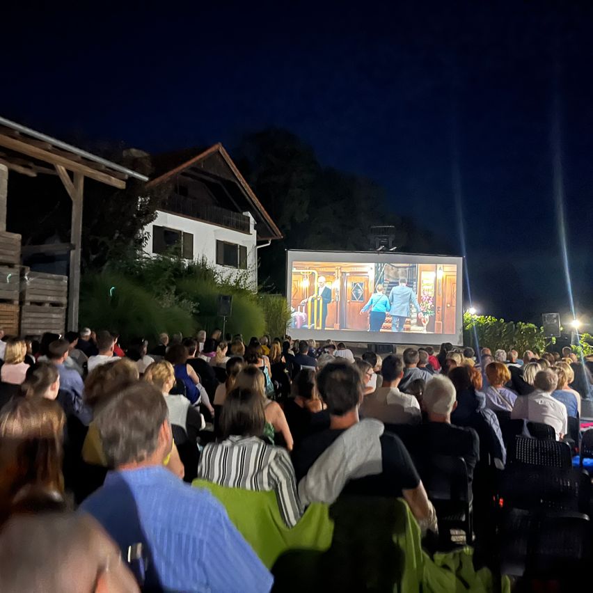 Eine nächtliche Freiluftkinovorführung. Eine große Menschenmenge ist versammelt und sieht einen auf einer Leinwand projizierten Film. Die Umgebung umfasst ein Haus im Hintergrund und Scheinwerfer im Vordergrund.