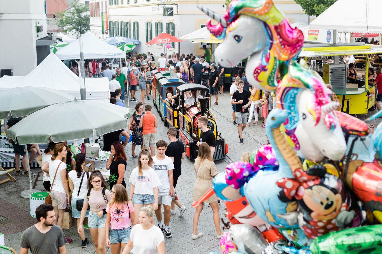 Eine Menschenmenge geht auf einer belebten Straße mit bunten Luftballons und einer Mini-Lokomotive. Zelte und Geschäfte sind an den Seiten aufgebaut. Einige Leute fahren mit der Lok, während andere gehen.