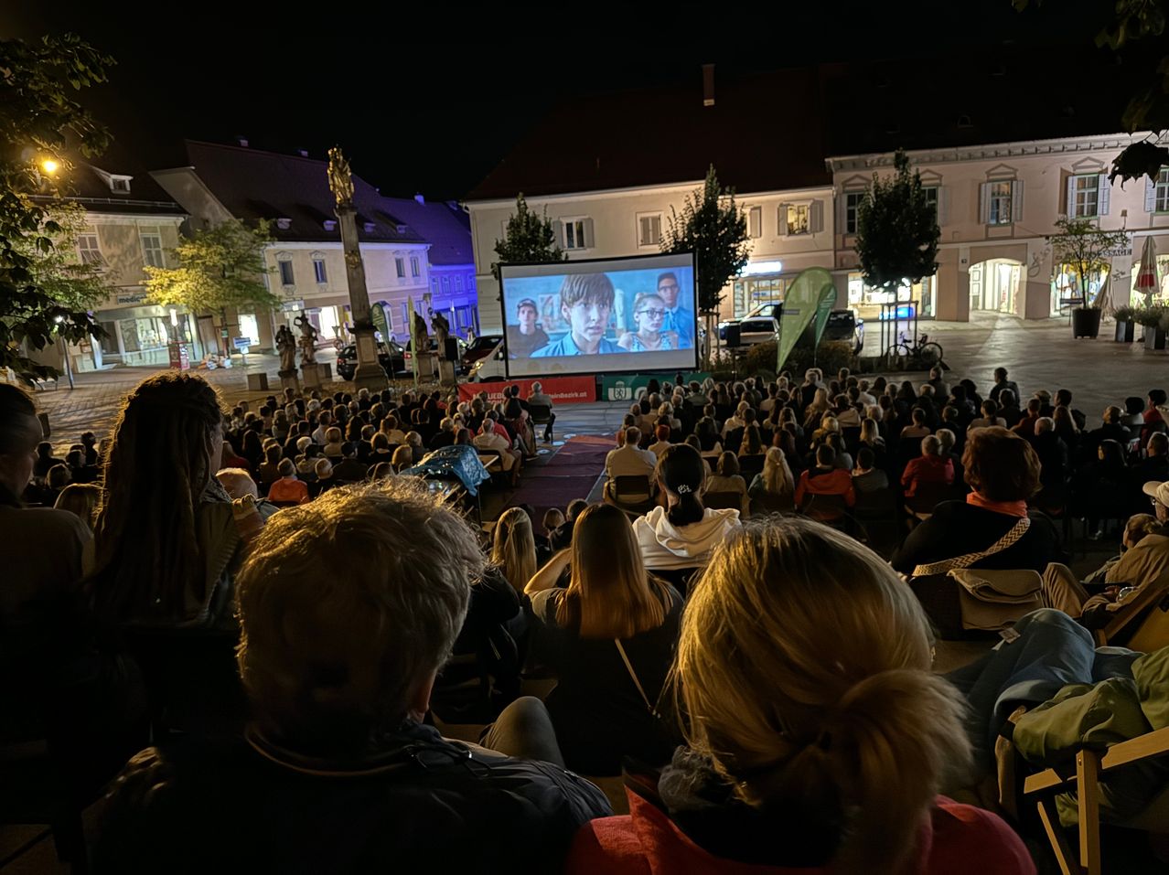 Eine nächtliche Freiluftkinovorführung findet auf einem Stadtplatz statt, wobei eine große Menschenmenge einen Film auf einer Projektionsleinwand ansieht. Die Leute sitzen auf Stühlen und Bänken.