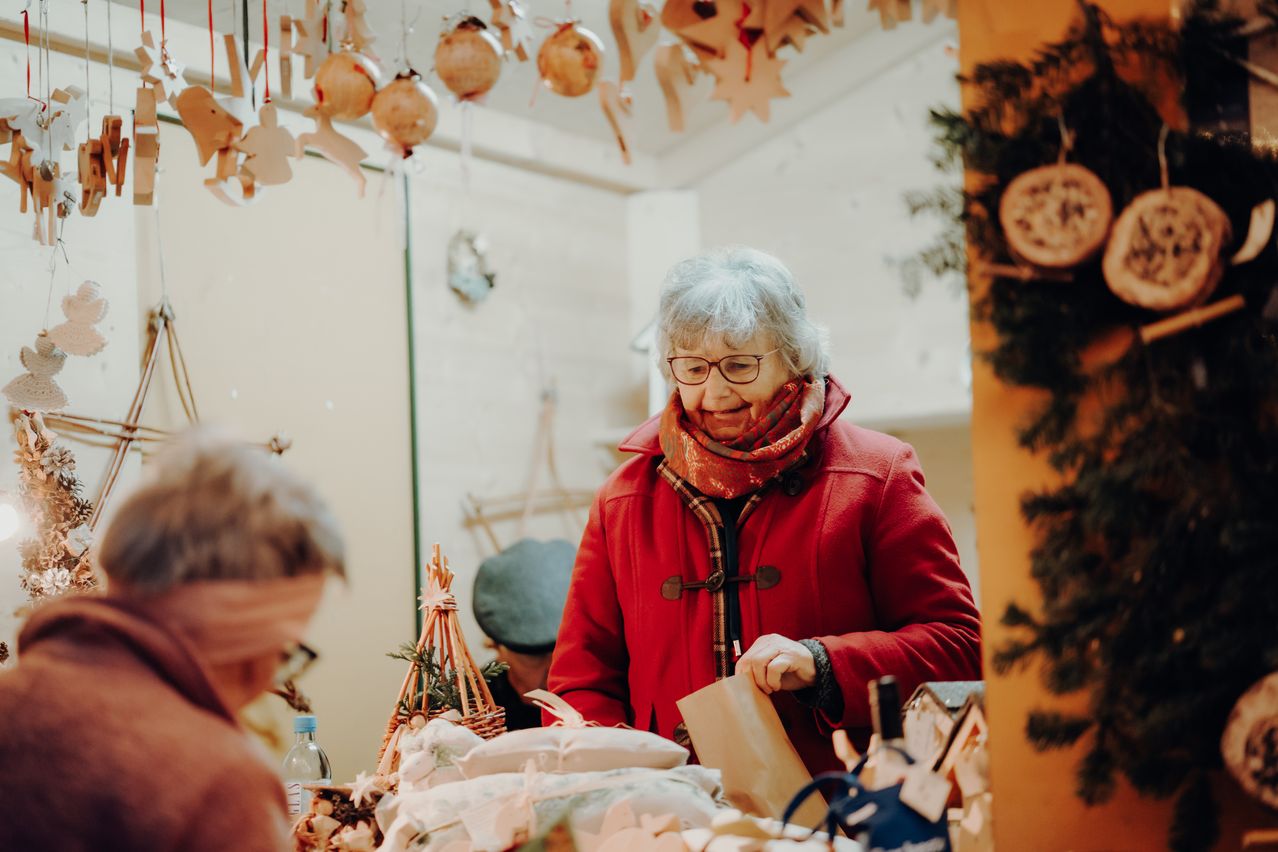 Eine ältere Frau in einem roten Mantel und einem Schal steht lächelnd am Tisch und hält einen braunen Papierbeutel. Der Tisch ist mit verschiedenen Gegenständen geschmückt, darunter ein Korb, eine Flasche und ein eingewickeltes Geschenk. Die Umgebung scheint ein festlicher, überdachter Markt zu sein.