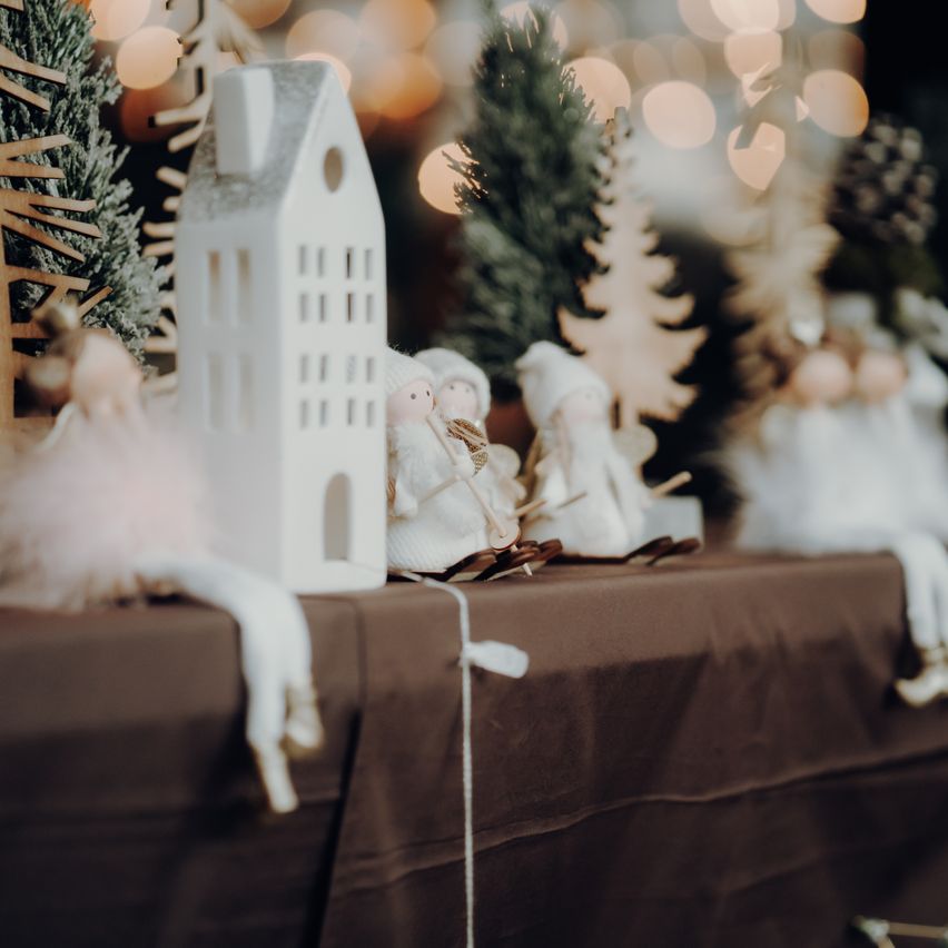 Eine weiße Hausfigur steht auf einem Tisch, umgeben von kleinen Weihnachtsbäumen und Engelfiguren, mit einem sanft verschwommenen Hintergrund.