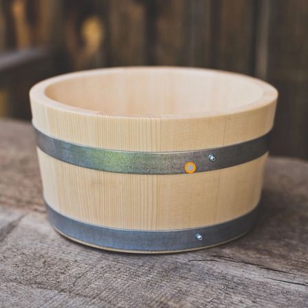 Ein holzfarbener Behälter mit einem silbernen Metallband in der Mitte und einem orangefarbenen Aufkleber an der Seite, auf einer Holzoberfläche platziert.