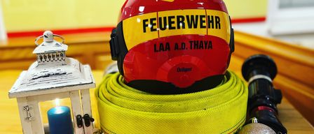 Ein Feuerwehrhelm wird auf einem gelben Schlauch mit Text ausgestellt. In der Nähe sind eine weiße Laterne, eine blaue Kerze und silberne Ornamente. Hinter dem Helm befindet sich ein roter Banner mit Text.