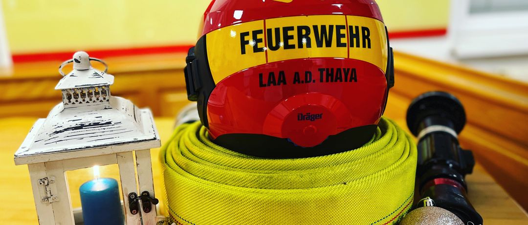 Ein Feuerwehrhelm wird auf einem gelben Schlauch mit Text ausgestellt. In der Nähe sind eine weiße Laterne, eine blaue Kerze und silberne Ornamente. Hinter dem Helm befindet sich ein roter Banner mit Text.