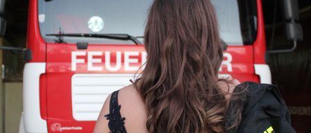 Eine Frau mit langen Haaren steht vor einem roten Feuerwehrauto, das mit 'LAA A.D.THAYA' beschriftet ist. Sie trägt ein schwarzes Kleid und eine Jacke mit gelbem und schwarzem reflektierendem Text.