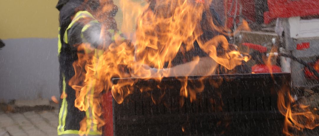 Ein Kind in voller Feuerwehrausrüstung steht neben einem brennenden Objekt, während ein anderer Feuerwehrmann Wasser darauf sprüht. Die Szene spielt sich im Freien auf einem Ziegelboden ab, mit einem Gebäude im Hintergrund.