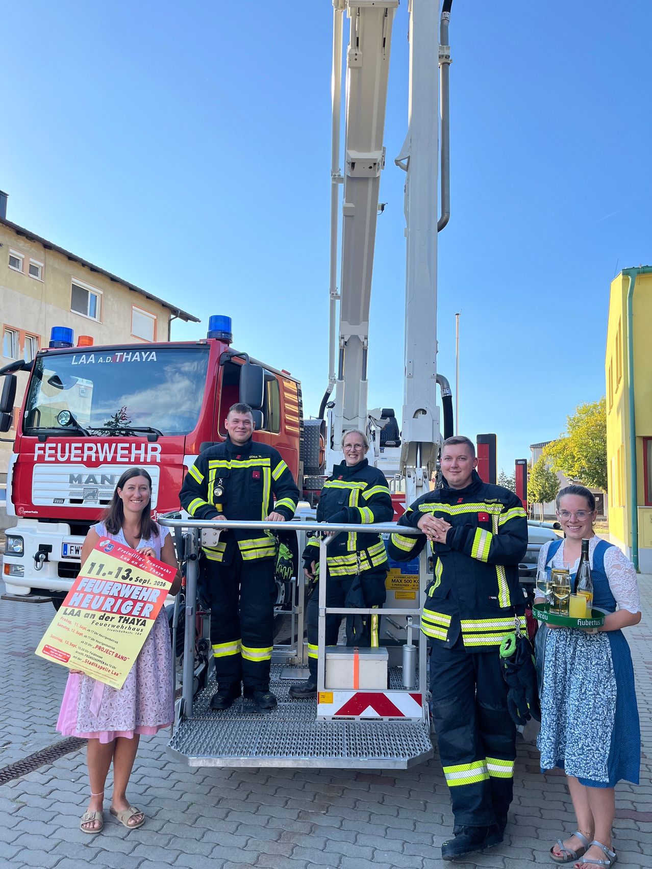 Eine Gruppe von Feuerwehrleuten posiert vor einem Feuerwehrauto und einer Leiter an einem sonnigen Tag. Eine Frau hält ein Schild, und zwei weitere stehen in der Nähe.