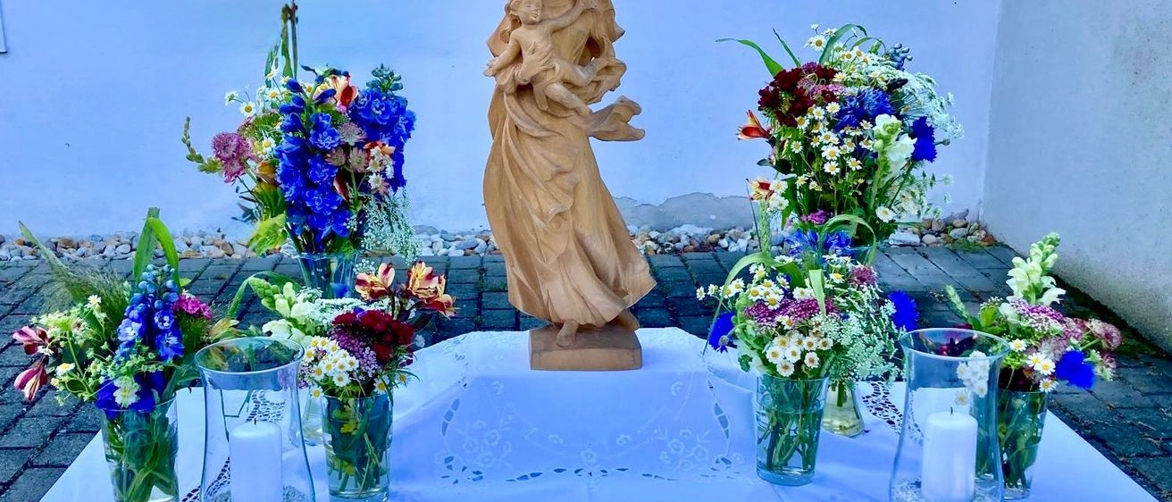 Eine Statue einer Frau, die ein Kind hält, steht auf einem Tisch, umgeben von Vasen mit Blumen, vor einem blauen Hintergrund.