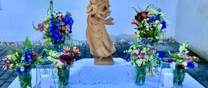 Eine Statue einer Frau, die ein Kind hält, steht auf einem Tisch, umgeben von Vasen mit Blumen, vor einem blauen Hintergrund.
